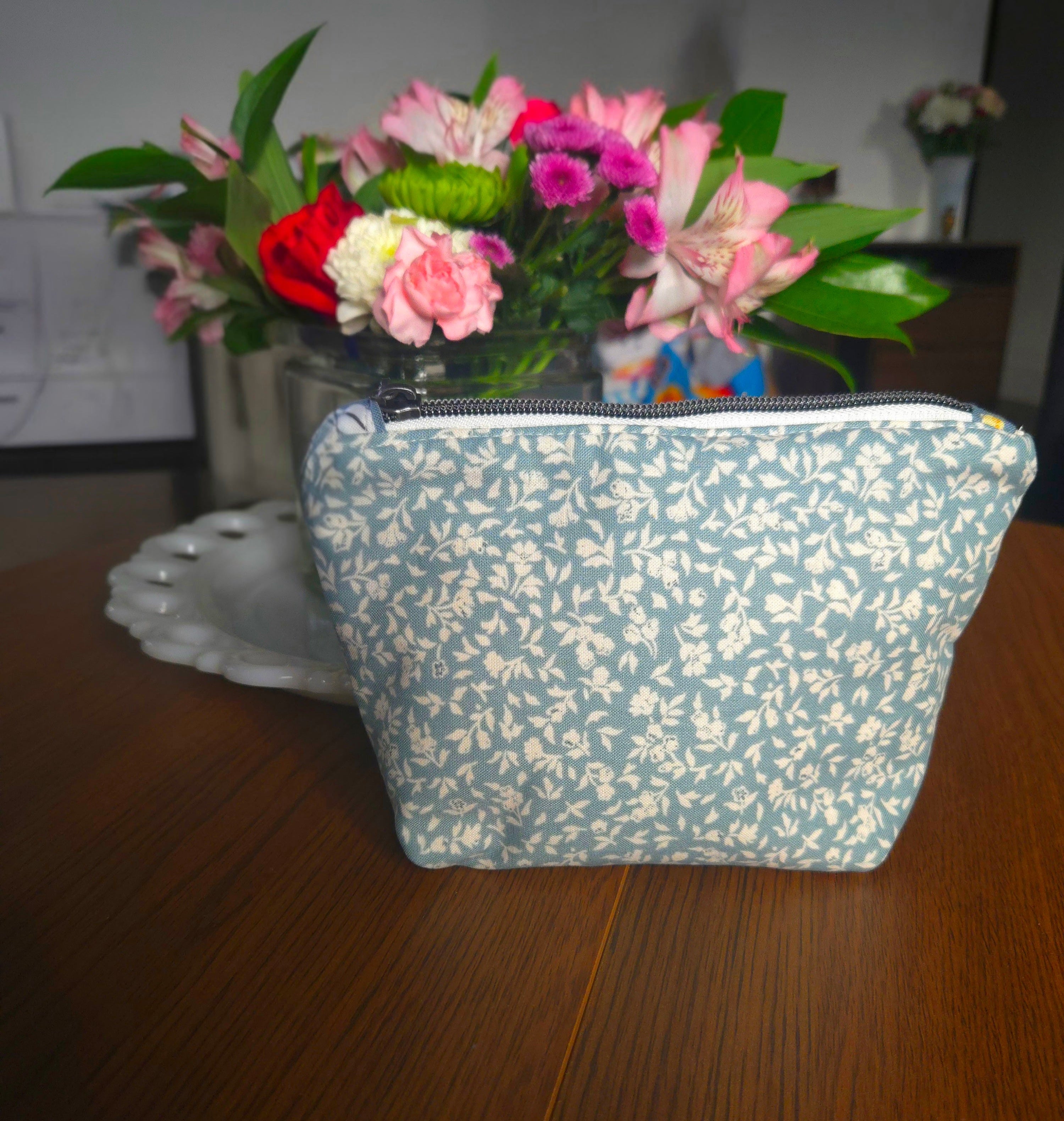 Floral-patterned pouch on a wooden surface with a vase of flowers in the background